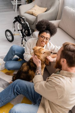Gülümseyen Afro-Amerikan kadın engelli köpek ve dövmeli erkek arkadaşının yanında oturma odasında oturuyor. 