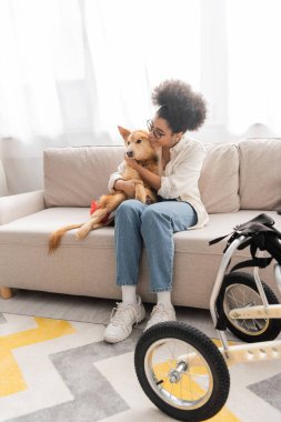 Pozitif Afro-Amerikalı kadın oturma odasındaki tekerlekli sandalyenin yanında engelli köpeğe sarılıyor. 