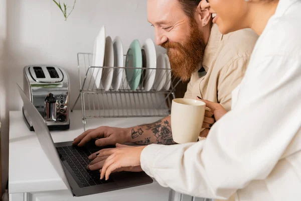 Smiling bearded man using laptop near african american girlfriend with coffee in kitchen