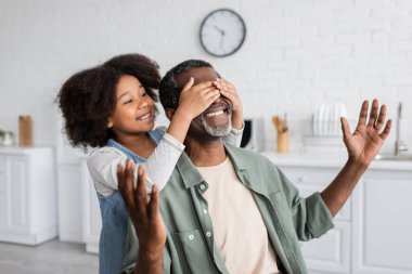 happy african american girl with curly hair covering eyes of smiling grandfather at home  clipart