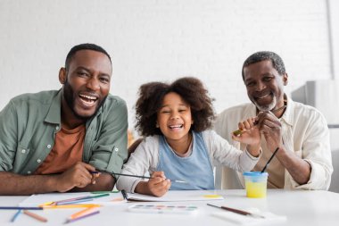 Smiling african american girl looking at camera near father and granddad while painting at home  clipart