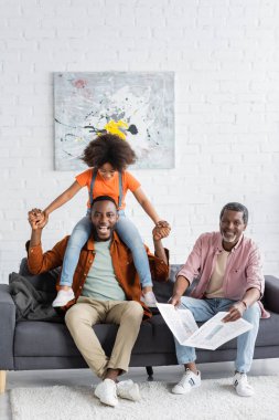 Smiling african american man having fun with daughter near dad holding newspaper at home  clipart