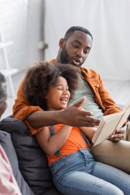 Afro-Amerikalı adam evde neşeli bir kız ve baba yanında kitap okuyor. 