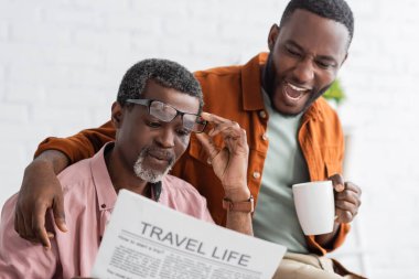 Excited african american man holding coffee cup while father reading newspaper at home  clipart