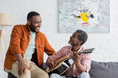 Olgun Afrikalı Amerikalı baba evdeki koltukta oğlunun yanında akustik gitar çalıyor. 