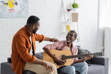 Gülümseyen Afrikalı Amerikalı adam oturma odasındaki kanepede yetişkin oğlunun yanında akustik gitar çalıyor. 