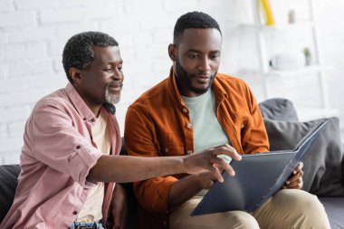 Gülümseyen Afrikalı Amerikalı adam evdeki koltukta oğlunun yanındaki fotoğraf albümüne bakıyor. 