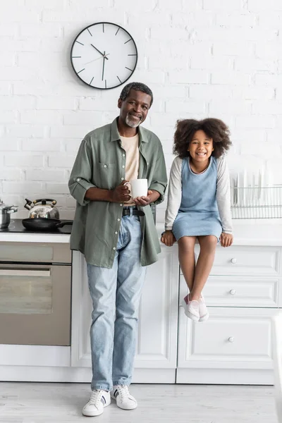 middle aged african american grandfather holding cup with coffee near joyful kid sitting on kitchen worktop