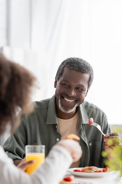 幸せなアフリカ系アメリカ人男性は新鮮なイチゴとフォークを持っていると朝食中に孫娘を見て — ストック写真