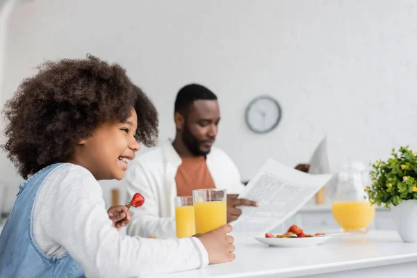 幸せなアフリカ系アメリカ人の女の子は朝食を食べながら父親はぼやけた背景で新聞を読んで — ストック写真