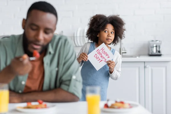 アフリカ系アメリカ人の女の子がお父さんの後ろに立っている間幸せな父親の日のレタリングでグリーティングカードを持っている — ストック写真