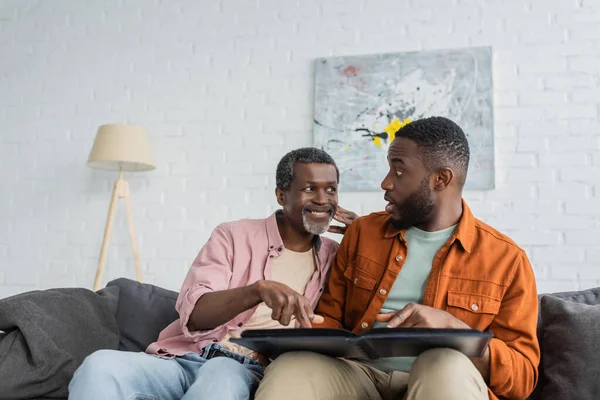 Smiling african american man pointing at photo album near shocked son at home