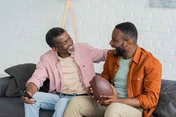 Pozitif Afro-Amerikan baba ve oğlu evde rugby topu ve uzaktan kumanda tutuyorlar. 