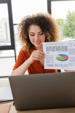 happy businesswoman with curly hair showing infographics during video call on laptop   clipart