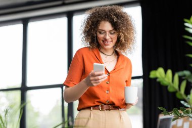 Gözlüklü neşeli iş kadını modern ofiste elinde kahve fincanı tutarken akıllı telefonuyla mesajlaşıyor. 