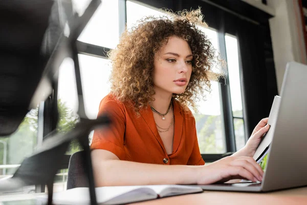 curly businesswoman holding folder and pen while using laptop in office