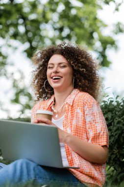joyful freelancer with curly hair holding paper cup and using laptop outdoors  clipart