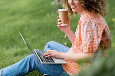 cropped view of cheerful freelancer with curly hair holding paper cup and using laptop while sitting on grass  clipart
