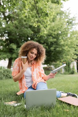 cheerful woman with curly hair holding newspaper and coffee to go while sitting on grass and looking at laptop  clipart