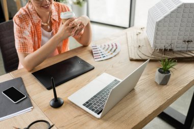 cropped view of happy architectural designer holding coffee to go near gadgets and residential house model in office clipart