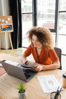 Neşeli ve kıvırcık bir kadın elinde klasörle masadaki çizelgelerin yanındaki dizüstü bilgisayarlara bakıyor. 