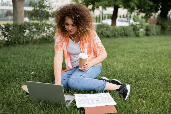 坐在草地上和使用笔记本电脑时 卷曲头发的年轻女子拿着咖啡要走 — 图库照片