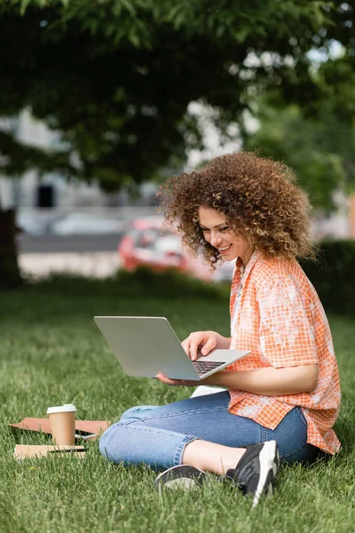 緑の公園でリモートで作業しながらラップトップを使用して巻き毛を持つ陽気なフリーランスの女性 — ストック写真