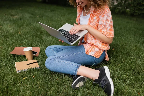 坐在靠近纸杯和笔记本的草坪上的快乐女人使用笔记本电脑的剪影 — 图库照片