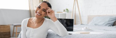 charming african american student smiling while sitting near laptop on bed, banner  clipart