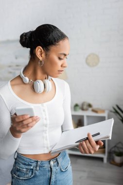 Boynunda kablosuz kulaklık olan Afro-Amerikan öğrenci akıllı telefon kullanıyor ve yatak odasında boş defter tutuyor. 