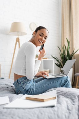 Gülümseyen Afrikalı Amerikalı öğrenci bilgisayarını kullanırken akıllı telefondan konuşuyor ve internetten öğreniyor. 