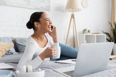 Neşeli Afro-Amerikan öğrenci elinde bir fincan kahveyle yatağında dizüstü bilgisayarla konuşuyor. 