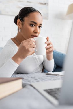 Güzel Afrikalı Amerikalı öğrenci elinde bir fincan kahveyle modern yatak odasında dizüstü bilgisayarda Webinar izliyor. 