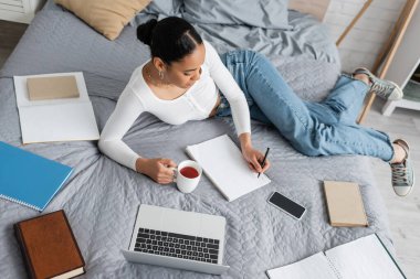 top view of african american student holding cup of tea and writing on notebook in bedroom  clipart