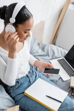 African american student in headphones holding cellphone near notebook on bed  clipart