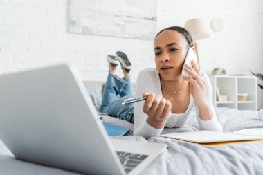 Afro-Amerikan öğrenci akıllı telefondan konuşuyor ve yatakta yatarken bulanık laptopa bakıyor. 