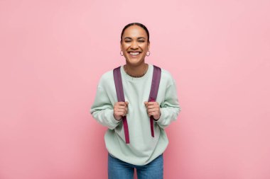 Smiling african american student holding backpack isolated on pink  clipart