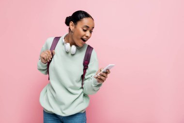 Heyecanlı Afro-Amerikan öğrenci pembe üzerine izole edilmiş akıllı telefon kullanıyor. 