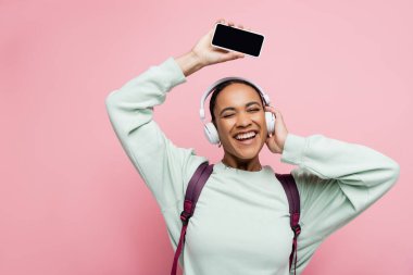 Kulaklıklı mutlu Afro-Amerikalı kadın elinde telefonuyla elinde boş ekranla pembe arka planda müzik dinliyor. 