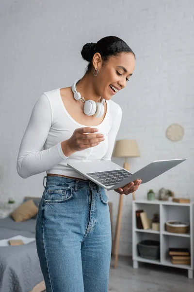 stock image excited african american student with wireless headphones on neck using laptop in modern bedroom 