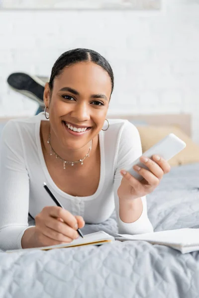 stock image Positive african american student holding cellphone and writing on notebook on bed 