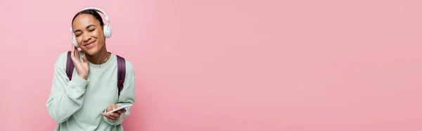 快乐的非洲裔美国女人 戴着无线耳机 手持智能手机 一边欣赏粉色横幅上的音乐 — 图库照片