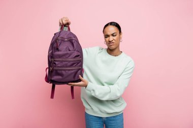 displeased african american student in sweatshirt holding purple backpack isolated on pink  clipart