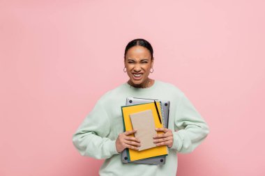 african american student in sweatshirt holding laptop and study supplies while grinning isolated on pink  clipart