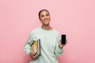 Sırıtan Afro-Amerikan öğrenci elinde çalışma malzemeleri ve pembe ekranlı akıllı telefon tutuyordu. 