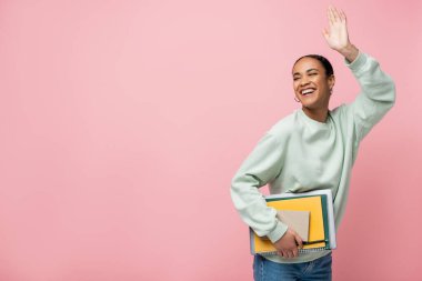 Eşofman altı giymiş neşeli Afro-Amerikan öğrenci elinde çalışma malzemeleri ve pembe üzerinde el sallıyor. 