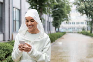 Kapüşonlu su geçirmez yağmurluk giyen mutlu Afro-Amerikalı kadın açık havada yağmurun altında dururken akıllı telefon kullanıyor. 