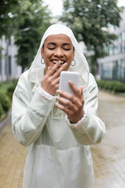 Kapüşonlu su geçirmez yağmurluk giyen neşeli Afro-Amerikalı kadın yağmurun altında dururken akıllı telefon kullanıyor. 