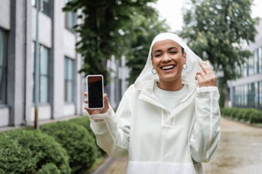 Kapüşonlu yağmurluk giyen ve elinde boş ekranlı akıllı telefon tutan mutlu Afro-Amerikan kadını. 