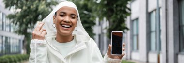 Suya dayanıklı yağmurluk giymiş neşeli Afro-Amerikalı kadın elinde beyaz ekranlı akıllı telefon, afiş 